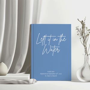 May include: A blue book with the text "Let it in the Water" in white script. The book also includes the name "Emily Jane", the date "September 19th, 2025", and "St. Mary's Church". The book is on a white surface.