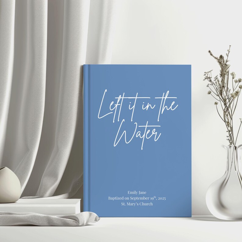 May include: A blue book with the text "Let it in the Water" in white script. The book also includes the name "Emily Jane", the date "September 19th, 2025", and "St. Mary's Church". The book is on a white surface.