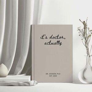 May include: A beige journal with the text "it's doctor, actually" in black script. Below the text is "DR. JACKSON, Ph.D EST. 2026". The journal is standing upright on a white surface, next to a vase with dried flowers.
