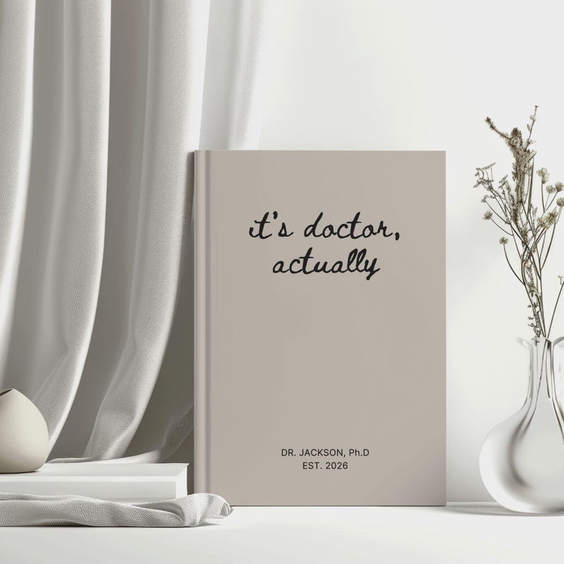 May include: A beige journal with the text "it's doctor, actually" in black script. Below the text is "DR. JACKSON, Ph.D EST. 2026". The journal is standing upright on a white surface, next to a vase with dried flowers.