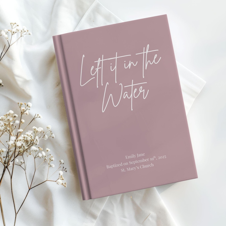 May include: A mauve-colored journal with the text "Lett it in the Water" in white script. The journal also includes the text "Emily Jane Baptized on September 10th, 2025 St. Mary's Church". The journal is on a white cloth.
