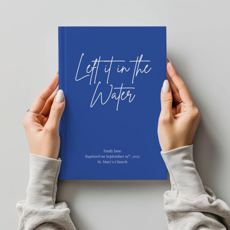 May include: A cobalt blue book with the text "Left it in the Water" in white script. The book also includes the name "Emily Jane", baptism date, and church name. The book is held by two hands.