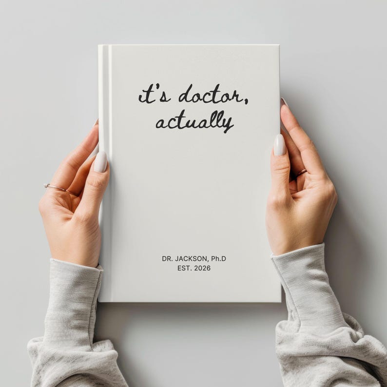 May include: A white hardcover journal with the text "it's doctor, actually" in black script. The journal also includes the text "DR. JACKSON, Ph.D EST. 2026". The journal is held by a person.
