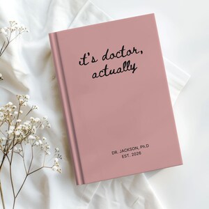 May include: A pink journal with the words "it's doctor, actually" in black script on the cover. The journal also has the text "DR. JACKSON, Ph.D EST. 2026". The journal is closed and on a white surface.