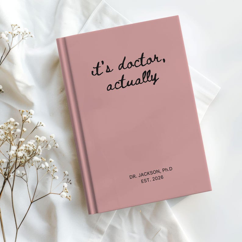 May include: A pink journal with the words "it's doctor, actually" in black script on the cover. The journal also has the text "DR. JACKSON, Ph.D EST. 2026". The journal is closed and on a white surface.