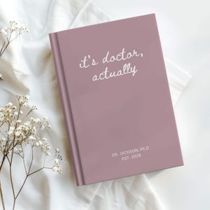 May include: A mauve journal with the text "it's doctor, actually" in white script. Below the text is "DR. JACKSON, Ph.D EST. 2026". The journal is on a white cloth with small white flowers.