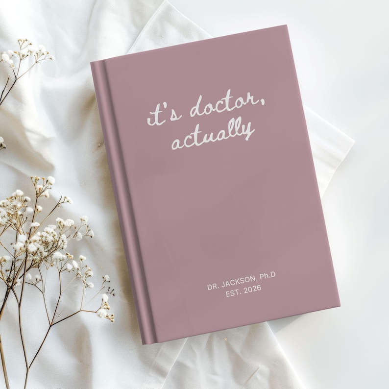 May include: A mauve journal with the text "it's doctor, actually" in white script. Below the text is "DR. JACKSON, Ph.D EST. 2026". The journal is on a white cloth with small white flowers.