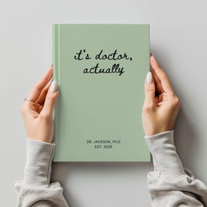 May include: A sage green notebook with the text "it's doctor, actually" in black script. The bottom of the notebook reads "DR. JACKSON, Ph.D EST. 2026". The notebook is held by two hands.