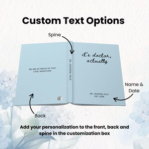 May include: Light blue notebook open to reveal the text "it's doctor, actually" on the right page. The left page reads "WE ARE SO PROUD OF YOU! LOVE, MOM & DAD". The spine reads "DR. JACKSON, Ph.D". The bottom of the right page reads "DR. JACKSON, Ph.D EST. 2026".