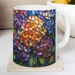 May include: White ceramic mug featuring a vibrant floral design. The artwork showcases a colorful arrangement of hydrangea blooms in shades of purple, blue, orange, and pink. The mug sits on a beige coaster.