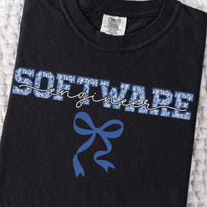 May include: Black t-shirt with the words "SOFTWARE ENGINEER" in blue floral print. The word "engineer" is written in a cursive font. A blue bow graphic is below the text. The shirt is folded on a white textured surface.