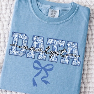 May include: A light blue t-shirt with the words "DATA analyst" in a white and blue floral design. A blue bow is below the text. The shirt is folded on a white textured surface.