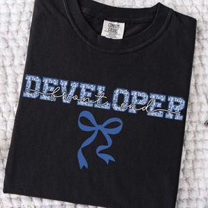 May include: Black t-shirt with the words "DEVELOPER front end" in blue floral print and a blue bow graphic. The shirt is folded on a white textured surface, with a star-shaped decoration and denim jeans visible.