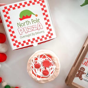 May include: A festive image featuring a pizza and a pizza box. The pizza box is white with a red checkered border and the words "North Pole Pizza Overnight Elf Delivery". The pizza has red sauce, white icing, and red pepperoni.