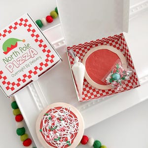 May include: A festive pizza cookie set with a red and white checkered pizza box that reads "North Pole Pizza Overnight Elf Delivery." The set includes a cookie decorated to look like a pizza, a piping bag, and sprinkles.