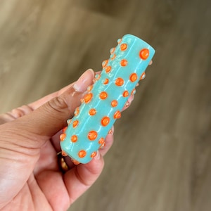 K&ouml;nnte beinhalten: Ein zylindrisches Glasobjekt in hellt&uuml;rkiser Farbe mit einer strukturierten Oberfl&auml;che, die mit zahlreichen erhabenen, leuchtend orangefarbenen kreisf&ouml;rmigen Punkten verziert ist, was ein taktiles und farbenfrohes Erscheinungsbild erzeugt.