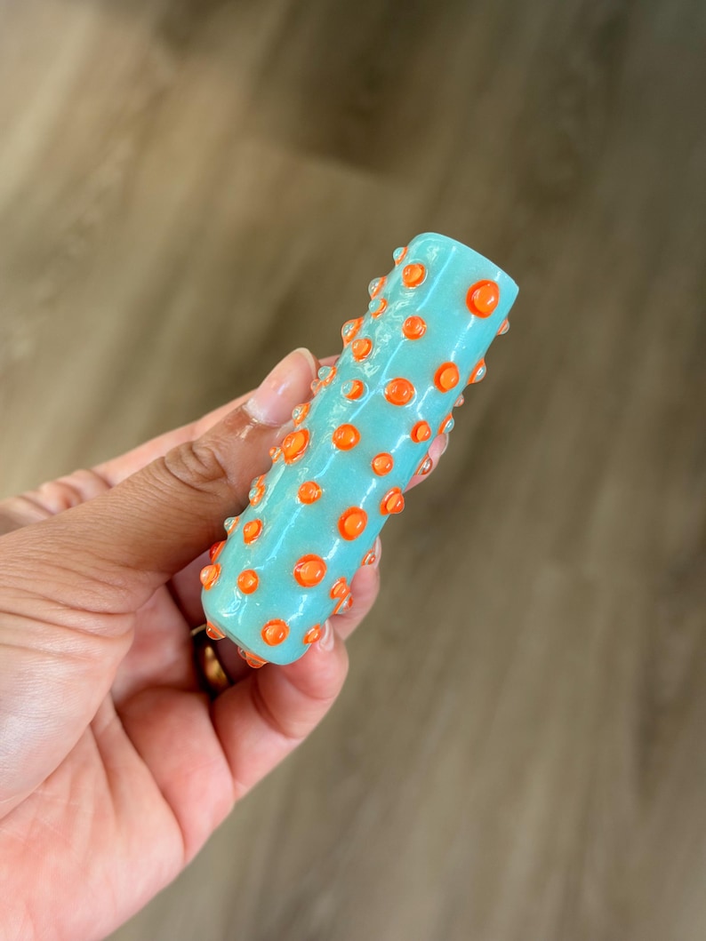 K&ouml;nnte beinhalten: Ein zylindrisches Glasobjekt in hellt&uuml;rkiser Farbe mit einer strukturierten Oberfl&auml;che, die mit zahlreichen erhabenen, leuchtend orangefarbenen kreisf&ouml;rmigen Punkten verziert ist, was ein taktiles und farbenfrohes Erscheinungsbild erzeugt.
