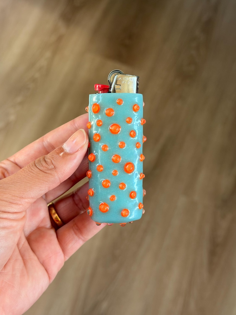 K&ouml;nnte beinhalten: Ein hellblaues Feuerzeug mit einer strukturierten Oberfl&auml;che, die mit kleinen, erhabenen orangefarbenen Punkten bedeckt ist. Das Feuerzeug hat einen silbernen Metallmechanismus mit einem roten Z&uuml;ndknopf f&uuml;r einen taktilen Griff.