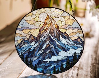May include: Round stained glass window art depicting a mountain scene with a sun and clouds. The mountain is brown and white, with blue and white clouds. The sky is yellow and blue. The window is framed in black and hangs from a chain.