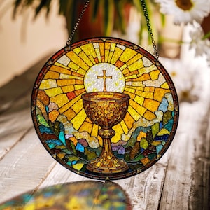 May include: Circular stained glass window depicting a religious scene. It features a chalice, a cross, and a sunburst design in yellow, orange, and brown hues. The window is framed with a dark border and suspended by a chain.
