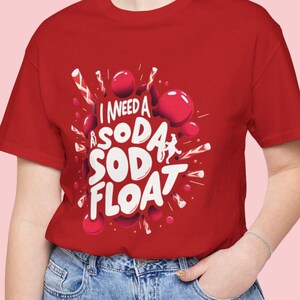 Może przedstawiać: Czerwony T-shirt z białym nadrukiem, na którym widnieje napis "Potrzebuję sodę z lodem" z czerwonymi i białymi ilustracjami bąbelków.