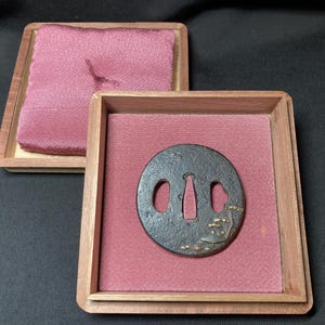 Puede incluir: Un tsuba de metal negro, una guarda decorativa para una espada japonesa, con detalles dorados. El tsuba se muestra en una caja de madera con un forro de terciopelo rosa.