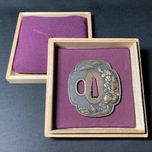 May include: An antique Japanese tsuba, a sword guard, presented in a wooden box with a purple fabric lining. The tsuba features a detailed relief design with a figure and landscape elements, showcasing traditional craftsmanship.
