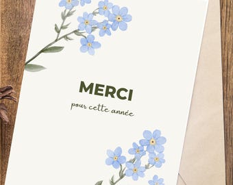 May include: A cream-coloured greeting card featuring blue watercolour forget-me-not flowers and the word "MERCI" in dark green. The phrase "pour cette année" is below the word "MERCI". The card is inside a light brown envelope, on a wooden surface.