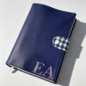 May include: A navy blue journal with a silver monogram of the letters "EA". The journal has a textured cover and a gingham patterned closure. A white ribbon bookmark is visible.