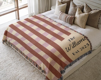 May include: A woven throw blanket with a striped pattern in shades of red, pink, and cream. The blanket is draped over a bed and features the text "The Williams Est 2025" in black script. The blanket has a fringed edge.