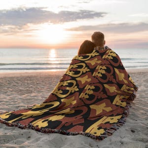 May include: A woven throw blanket featuring a repeating pattern of gold and dark red crown motifs on a black background. The blanket has a fringed edge and is draped over the shoulders of two individuals sitting on a sandy beach at sunset.