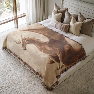 May include: A beige throw blanket featuring a detailed illustration of a brown dragon with outstretched wings. The blanket has a fringed edge with a mix of colors. The dragon is the focal point, set against a neutral background.