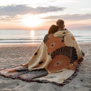 May include: A beige and brown floral patterned throw blanket with a fringed edge, draped over two people on a sandy beach. The blanket features large, stylized flower designs in shades of brown and cream. The background shows the ocean and a sunset.