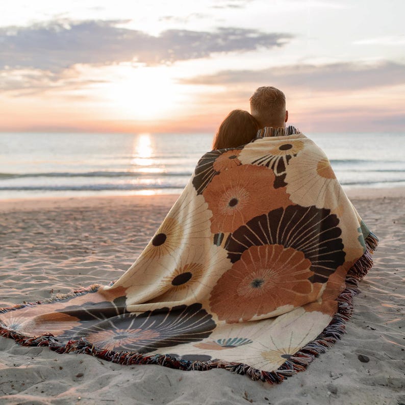 May include: A beige and brown floral patterned throw blanket with a fringed edge, draped over two people on a sandy beach. The blanket features large, stylized flower designs in shades of brown and cream. The background shows the ocean and a sunset.