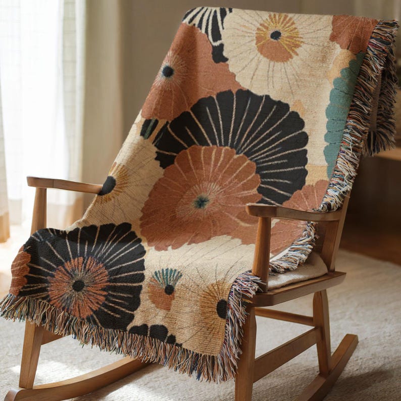 May include: A floral patterned throw blanket draped over a wooden rocking chair. The blanket features large, stylized flower designs in shades of brown, black, and beige, with fringed edges. The chair is made of light-colored wood.