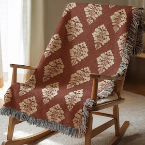 May include: A woven throw blanket with a floral pattern in cream and rust red is draped over a wooden rocking chair. The blanket has a fringed edge in shades of gray, blue, and cream. The floral design is repeated across the blanket's surface.