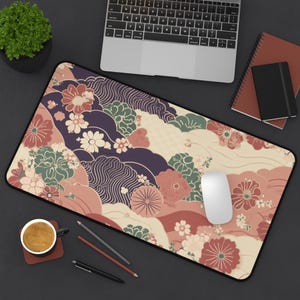 Puede incluir: Alfombrilla de escritorio rectangular con un estampado floral y de olas en tonos rosa, crema, verde y morado. La alfombrilla tiene un borde negro y está diseñada para un teclado y un ratón. Se ven un portátil, un ratón y artículos de papelería.