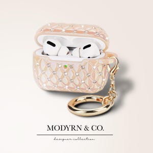 May include: A pink and gold Airpods case with a diamond pattern and a gold chain and ring. The case is open and the Airpods are inside. The text "MODYRN & CO. designer collection" is at the bottom of the image.