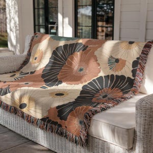 May include: A woven throw blanket with a floral pattern in shades of beige, brown, and black, draped over a white wicker sofa. The blanket features a fringed edge and a repeating design of stylized flowers.