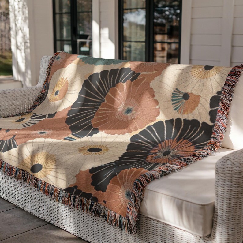 May include: A woven throw blanket with a floral pattern in shades of beige, brown, and black, draped over a white wicker sofa. The blanket features a fringed edge and a repeating design of stylized flowers.