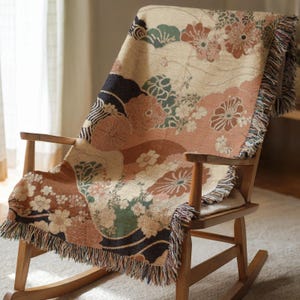 Puede incluir: Una manta tejida con un estampado floral en tonos marrones, rosas y verdes está sobre una mecedora de madera. La manta tiene un borde con flecos y un fondo beige. La silla es de madera clara.