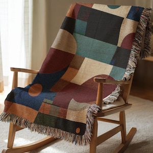 May include: A woven throw blanket with a geometric pattern in shades of blue, burgundy, tan, and green, draped over a wooden rocking chair. The blanket features a fringed edge.