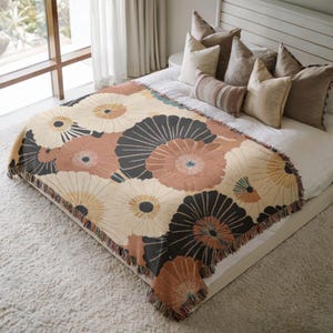 May include: A floral patterned throw blanket in cream, peach, and black, with a fringed edge, is draped over a bed. The design features large, stylized flower shapes. The blanket is on a white bed with several pillows.