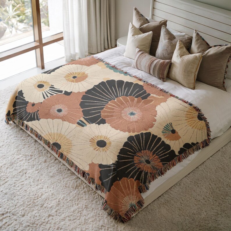 May include: A floral patterned throw blanket in cream, peach, and black, with a fringed edge, is draped over a bed. The design features large, stylized flower shapes. The blanket is on a white bed with several pillows.
