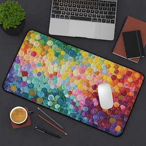 May include: A rectangular mouse pad with a vibrant, abstract design of colorful circles in shades of blue, green, yellow, orange, and purple. A white computer mouse sits on the pad. A laptop, notebooks, and a cup of coffee are also visible.