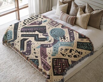 May include: A beige throw blanket with a geometric pattern in shades of blue, purple, and black. The blanket features a fringe trim and is displayed on a bed with pillows. The design includes abstract shapes and dotted details.