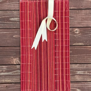 May include: A rectangular fabric pouch featuring a vertical striped pattern in shades of red, orange, and dark maroon. The pouch is secured with a cream-colored ribbon tied in a loop at the top center, with two loose ends hanging down the front.