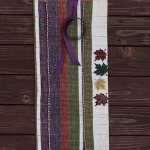 May include: A rectangular, woven placemat with vertical stripes in purple, red, green, and white. A purple ribbon is tied near the top. The right side features embroidered fall leaves in red, green, and gold.