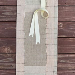 May include: A rectangular fabric organiser featuring a central beige panel flanked by light pink grid-patterned side sections. The item includes a cream-coloured ribbon loop at the top for hanging and visible stitching along the edges and grid lines.