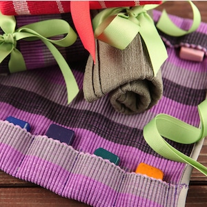May include: A purple and gray striped fabric roll-up case with multiple slots for holding small items, such as crayons or markers. The case is decorated with green ribbon and a rolled up gray fabric item.